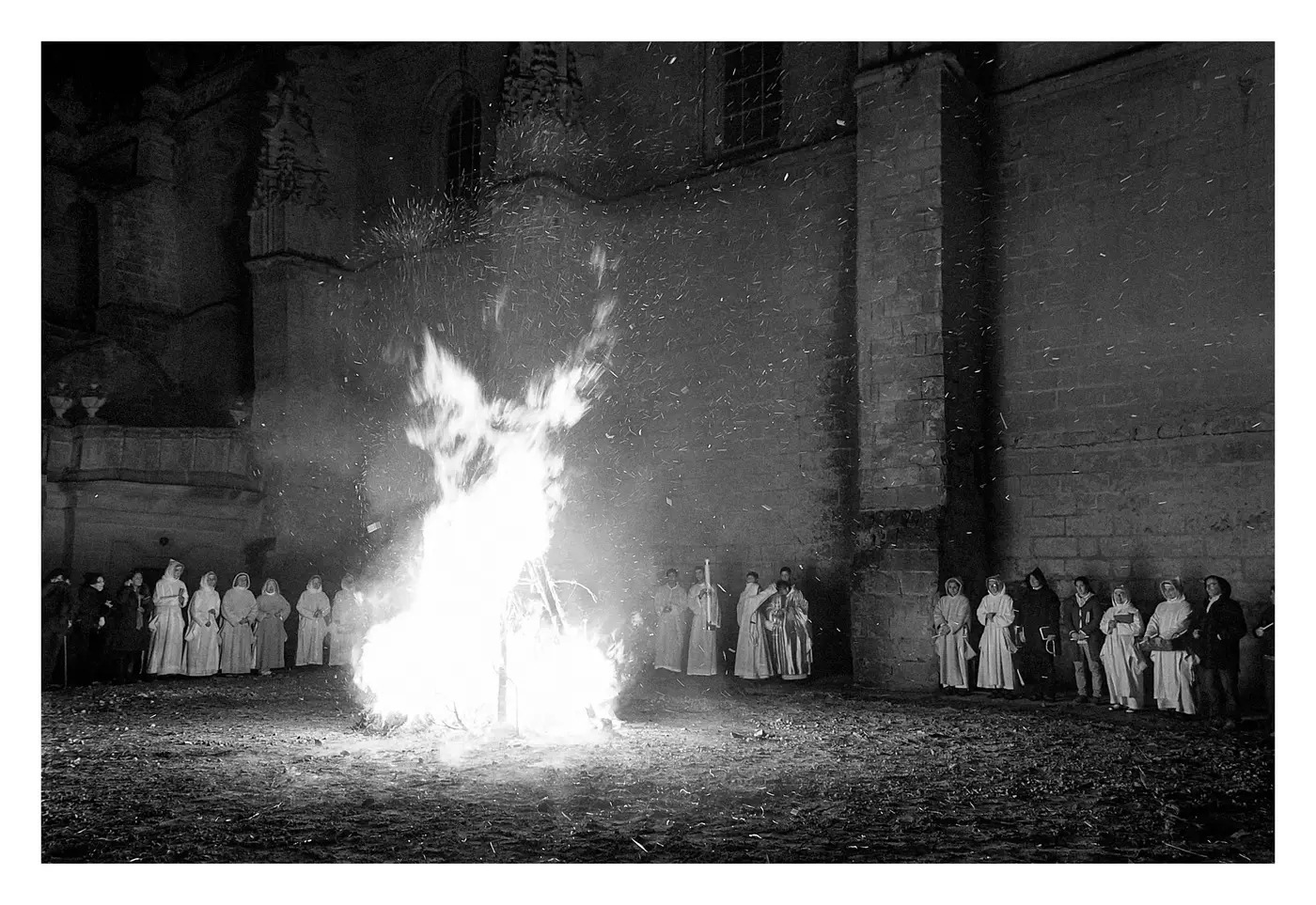 Gran hoguera ardiendo de noche frente al muro del monasterio, rodeada por la comunidad con hábitos blancos