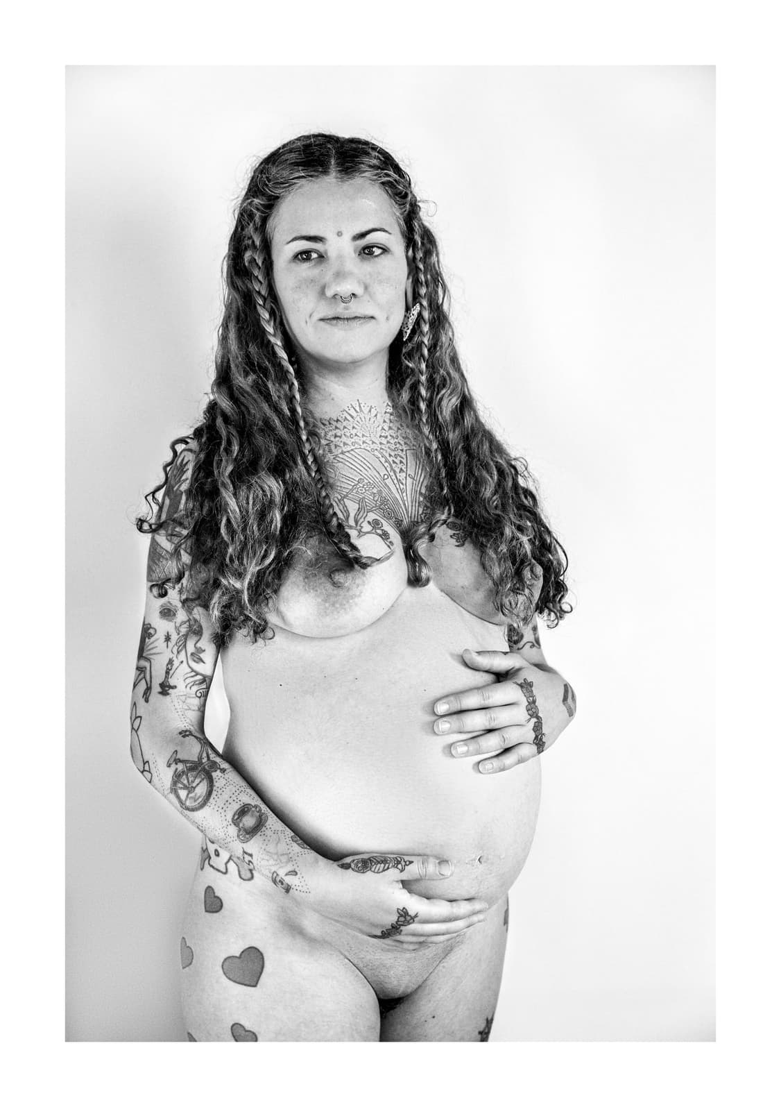Retrato de una mujer embarazada desnuda y tatuada, de pie, sosteniéndose el vientre con las dos manos