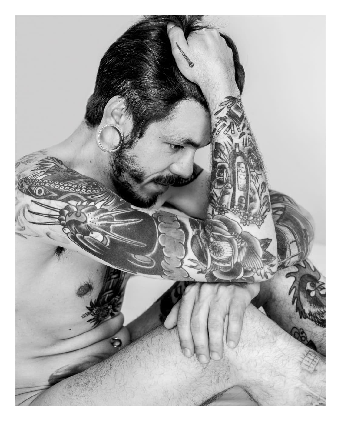 Hombre desnudo y tatuado sentado, con una mano en el pelo y la mirada baja