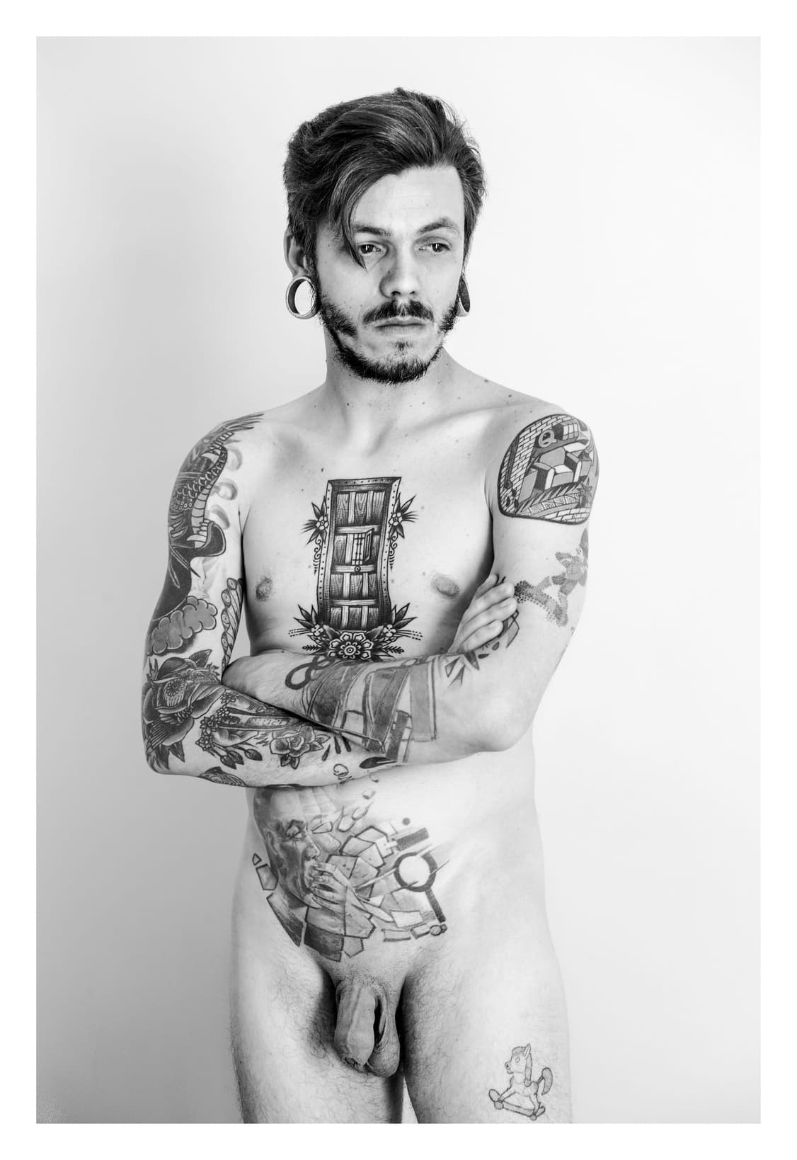 Retrato de cuerpo entero de un hombre desnudo y tatuado, de pie y con los brazos cruzados