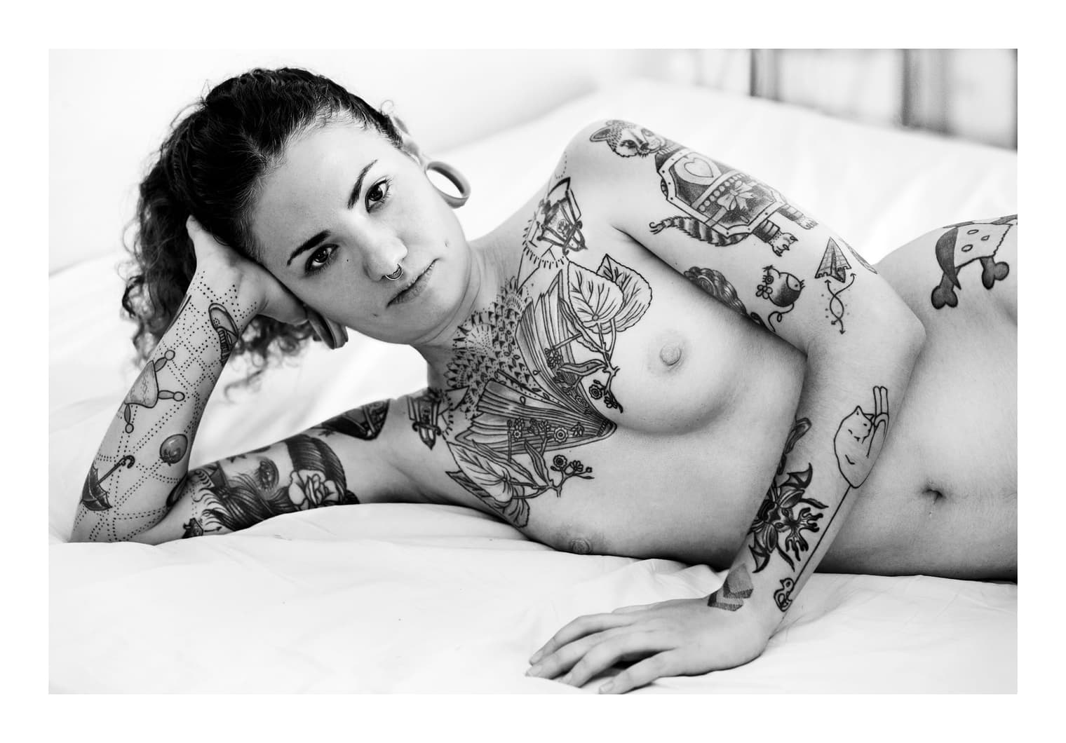 Mujer desnuda y tatuada tendida en una cama, con la cabeza apoyada en la mano, mirando a cámara
